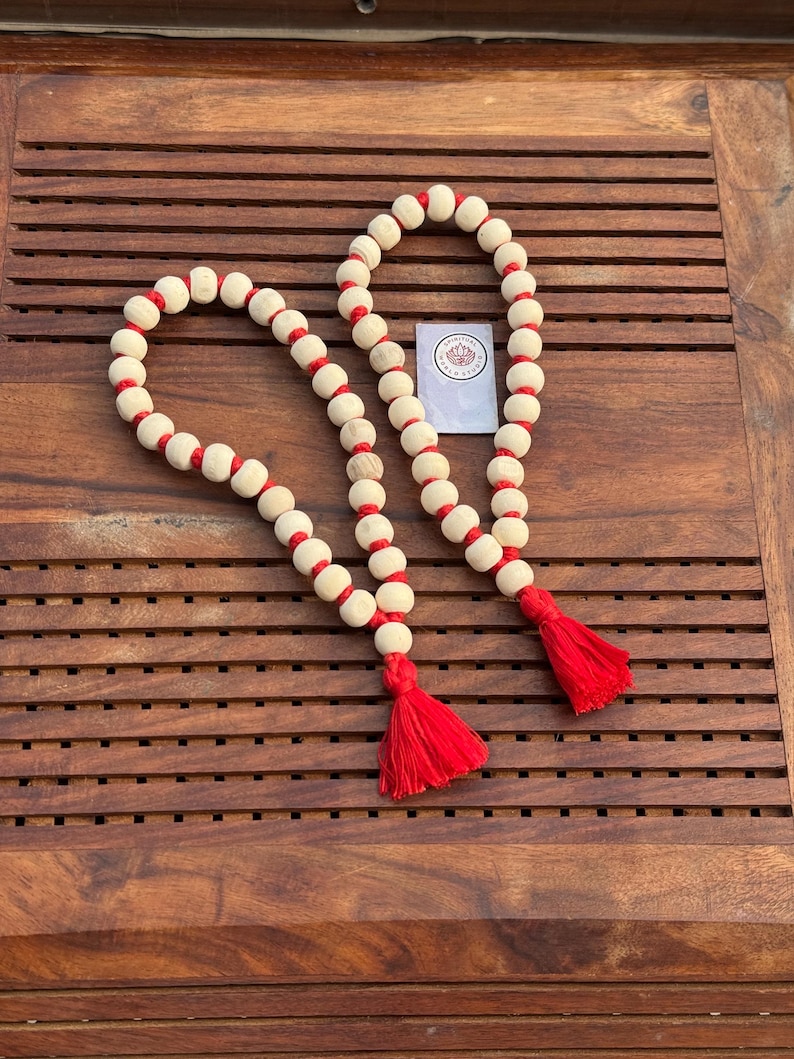 May include: Two wooden bead garlands with red string accents and red tassels. The garlands are on a wooden surface.
