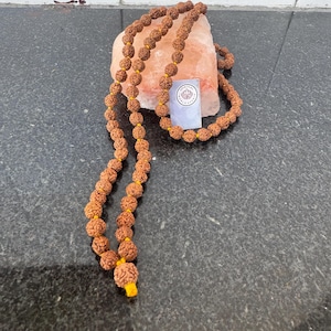 Könnte beinhalten: Eine braune Mala-Perlenkette mit gelbem Faden und einem Quaste. Die Kette besteht aus Rudraksha-Perlen und liegt auf einem rosa Himalaya-Salzstein.