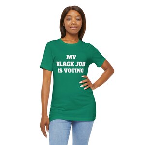 Könnte beinhalten: Ein grünes T-Shirt mit dem weißen Text "MY BLACK JOB IS VOTING". Die Person trägt eine blaue Jeans. Das Bild zeigt eine Person mit dunklen Haaren.