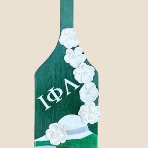 May include: A green wooden paddle decorated with white flowers and a painted image of a woman wearing a hat. The paddle has the Greek letters ΙΦΛ in white. A green ribbon is attached at the top.