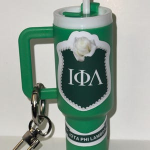 Könnte beinhalten: Grüner Reisebecher mit weißem Deckel und Strohhalm. Der Becher hat ein weißes und grünes Emblem mit einer Rose und den griechischen Buchstaben IΦΛ. Ein silberner Schlüsselanhänger ist am Griff befestigt. Der Text "IOTA PHI LAMBDA" ist auf dem Becher aufgedruckt.