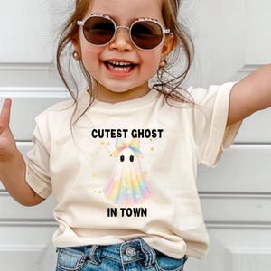 May include: A child wearing a cream-colored t-shirt with the text "Cutest Ghost in Town" and a colorful ghost graphic. The child is also wearing blue denim shorts and sunglasses. The child is smiling and giving a peace sign.