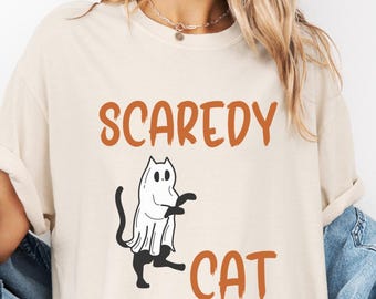 May include: Cream-colored t-shirt with the words "Scaredy Cat" in orange. A black cat wearing a ghost costume is in the center. The shirt has short sleeves and a relaxed fit, perfect for Halloween.