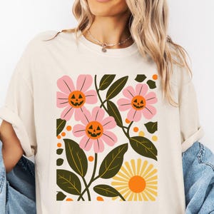 May include: Cream-colored t-shirt featuring a floral design with pink flowers, green leaves, and a yellow sun. The flowers have jack-o'-lantern faces, creating a Halloween theme. The shirt has a relaxed fit.