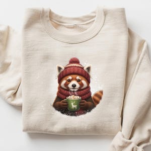 May include: A beige sweatshirt featuring a graphic of a red panda wearing a red knit hat and scarf, holding a green mug with a deer design. The red panda is surrounded by a snowy background. The sweatshirt is folded on a white surface.