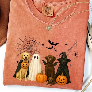 May include: Coral-colored t-shirt featuring a Halloween-themed graphic. The design includes illustrations of dogs dressed as a ghost, a witch, and a vampire, along with pumpkins, bats, and a spiderweb. The shirt has a crew neck and short sleeves.