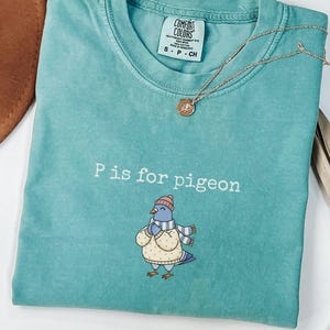 May include: A teal Comfort Colors t-shirt with the text "P is for pigeon" and an illustration of a pigeon wearing a hat and scarf. A gold necklace with a pendant is draped across the shirt.