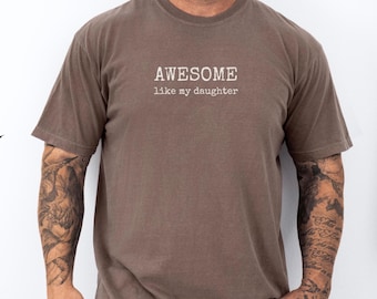May include: A brown t-shirt with the text "AWESOME like my daughter" in white. The shirt has short sleeves and a crew neck. The person wearing the shirt has tattoos on their arms.