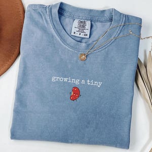 May include: A light blue t-shirt with the text "growing a tiny" and a small red bean illustration. The shirt has a distressed look and is made of a soft, comfortable fabric.