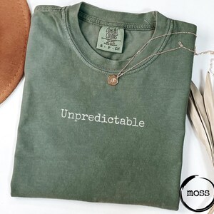 May include: An olive green t-shirt with the word "Unpredictable" in white script. A gold necklace with a pendant rests on the shirt. The shirt is folded and has a tag that says "Comfort Colors".
