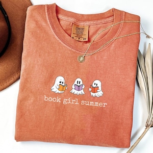 May include: An orange Comfort Colors t-shirt with a graphic of three cartoon ghosts reading books. The text "book girl summer" is printed below the ghosts. A gold necklace with a pendant is draped on the shirt.