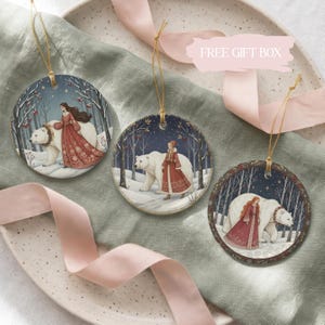 May include: Three round ceramic ornaments featuring illustrations of a person in a long red dress walking alongside a large white polar bear in a snowy forest setting. Each ornament hangs from a thin gold string and displays a unique winter scene.