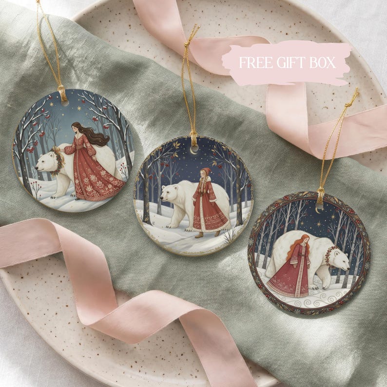 May include: Three round ceramic ornaments featuring illustrations of a person in a long red dress walking alongside a large white polar bear in a snowy forest setting. Each ornament hangs from a thin gold string and displays a unique winter scene.