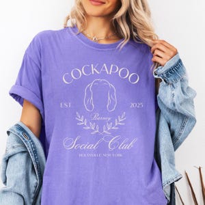 May include: Purple t-shirt with a white graphic design that says "Cockapoo Social Club" with a stylized image of a dog and the text "Est. 2025" and "Housville, New York".