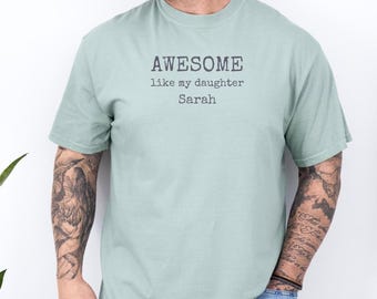 May include: A light green t-shirt with the text "AWESOME like my daughter Sarah" printed in black. The shirt has short sleeves and a crew neck. The person wearing the shirt has tattoos on both arms.
