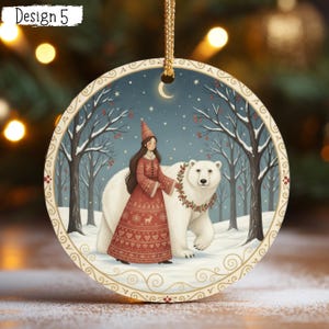 May include: A round ceramic ornament featuring a winter scene with a person in a red patterned dress and a pointed hat walking beside a large white polar bear wearing a holly wreath. The background shows snowy trees, a crescent moon, and stars.