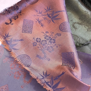May include: Close-up of several pieces of fabric with an Asian-inspired design. The fabric has a sheen and features a pattern of bamboo, flowers, and geometric shapes in shades of purple, bronze, and green.