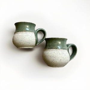 May include: Two handmade ceramic mugs. The top half of each mug is a glossy, muted green, while the bottom half is speckled white. Each mug has a rounded body and a curved handle, set against a white background.