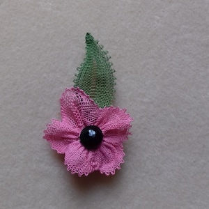 Puede incluir: Una flor de crochet rosa con un botón negro en el centro y una hoja de crochet verde.