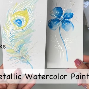 Op de afbeelding: Twee bladwijzers met aquarelverf. De ene bladwijzer heeft een blauwe en gele pauwenveer. De andere bladwijzer heeft een blauwe bloem met zilveren stelen en bladeren. De tekst "Bookmarks with Metallic Watercolor Paints" is zichtbaar onderaan de afbeelding.