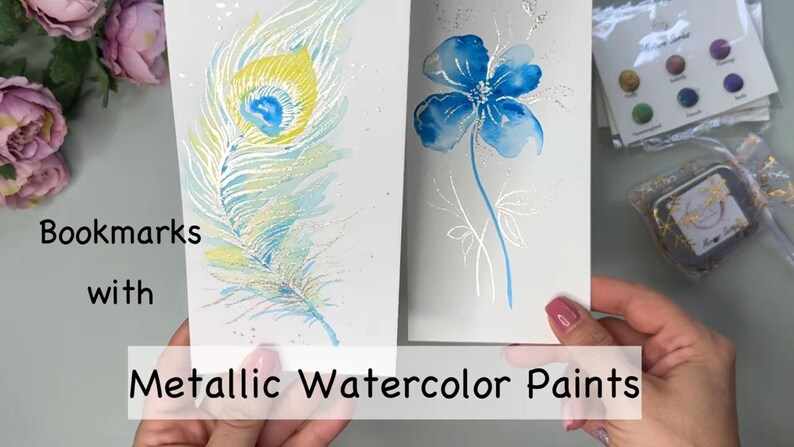 Op de afbeelding: Twee bladwijzers met aquarelverf. De ene bladwijzer heeft een blauwe en gele pauwenveer. De andere bladwijzer heeft een blauwe bloem met zilveren stelen en bladeren. De tekst "Bookmarks with Metallic Watercolor Paints" is zichtbaar onderaan de afbeelding.