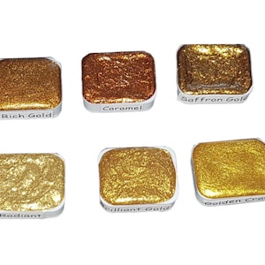 May include: Six watercolor paint pans in a silver tray. The pans are filled with metallic gold, copper, and yellow paint. The labels on the pans read: Rich Gold, Caramel, Saffron Gold, Radiant, Brilliant Gold, and Golden Crocus.