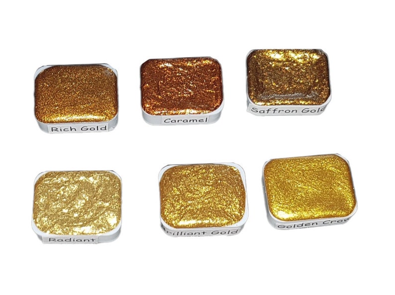 May include: Six watercolor paint pans in a silver tray. The pans are filled with metallic gold, copper, and yellow paint. The labels on the pans read: Rich Gold, Caramel, Saffron Gold, Radiant, Brilliant Gold, and Golden Crocus.