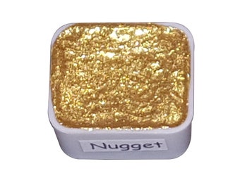 Puede incluir: Un pequeño recipiente cuadrado de pintura acuarela dorada metálica. El recipiente es blanco con una etiqueta negra que dice "Nugget".