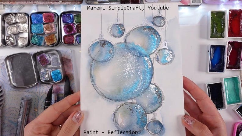 K&ouml;nnte beinhalten: Ein Aquarellbild von blauen und silbernen Weihnachtskugeln mit Glitzer. Das Bild ist auf wei&szlig;em Hintergrund und hat den Text "Paint - Reflection" unten.