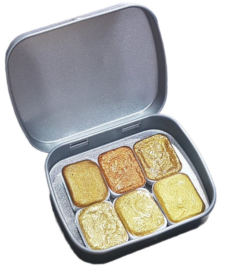 May include: A silver metal tin with six pans of metallic watercolor paint. The pans are arranged in two rows of three. The colors are gold, copper, and bronze.