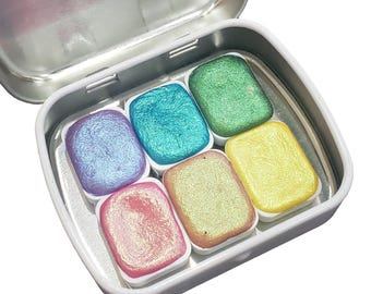 May include: A set of six metallic watercolor paints in a tin case. The colors are purple, teal, green, yellow, pink, and gold. Each paint pan has a shimmery finish.