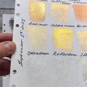 May include: A white card with swatches of gold paint. The swatches are labeled "Theol. 67 squs", "Saftrefold", "Caramel", "Radiant", "Golden crown", "Brilliant Gold", "Splendour", "Reflection", and "Illusion". The card also has the text "September 19' 10:34" written on it.