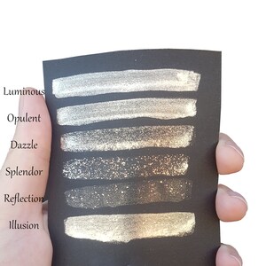 K&ouml;nnte beinhalten: Sechs Farbmuster aus Metallic-Goldfarbe auf einer schwarzen Karte. Die Farbmuster sind von oben nach unten beschriftet: Luminous, Opulent, Dazzle, Splendor, Reflection, Illusion.