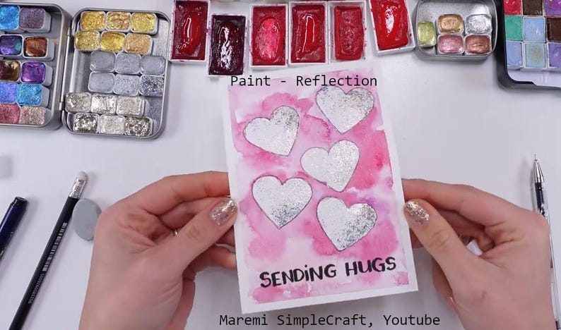 K&ouml;nnte beinhalten: Eine rosa Aquarellkarte mit f&uuml;nf silbernen Glitzerherzen und dem Text "Sending Hugs" in Schwarz. Die Karte ist signiert mit "Maremi SimpleCraft, Youtube".