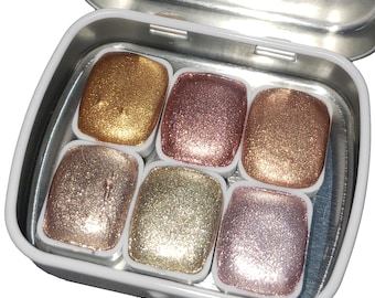 May include: A tin of six metallic watercolor paints in a variety of gold, rose gold, and silver shades. The paints are in a compact tin with a white lid.