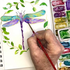 May include: A hand paints a watercolor dragonfly with a fine brush. The dragonfly is surrounded by a wreath of green leaves. The artist is using a palette of watercolors with a variety of colors.