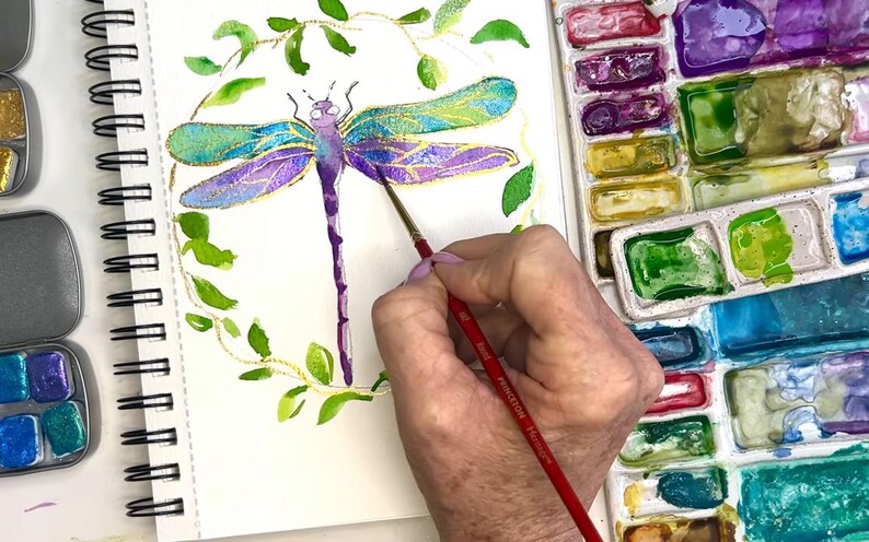 May include: A hand paints a watercolor dragonfly with a fine brush. The dragonfly is surrounded by a wreath of green leaves. The artist is using a palette of watercolors with a variety of colors.
