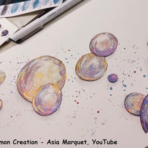 May include: A white sheet of paper with a watercolor painting of iridescent bubbles. The bubbles are painted in shades of pink, purple, blue, and yellow. The background is a light blue with white splatters.