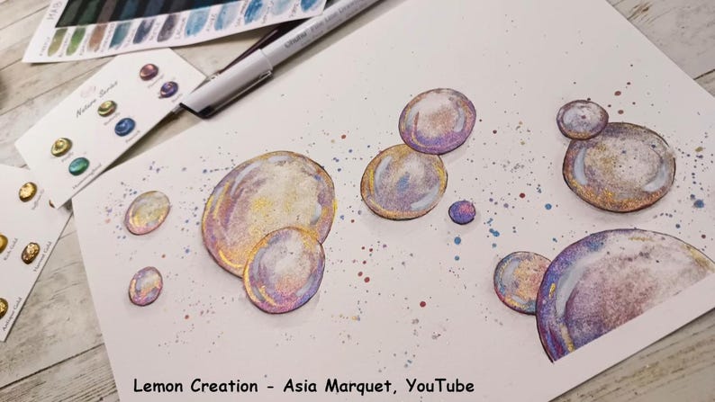 May include: A white sheet of paper with a watercolor painting of iridescent bubbles. The bubbles are painted in shades of pink, purple, blue, and yellow. The background is a light blue with white splatters.