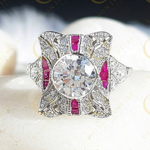 Op de afbeelding: Een vintage-stijl verlovingsring met een grote, ronde, heldere middelste steen omringd door kleinere diamanten en rode robijnen. De ring is gezet in een witte metalen band.