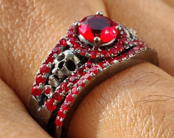May include: A silver ring with a large red gemstone surrounded by smaller red gemstones. The ring is set with a skull design and has a black band.
