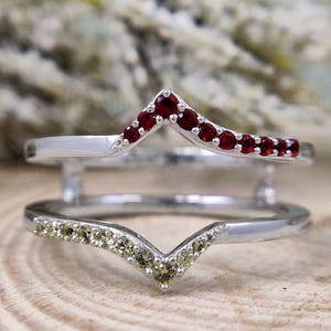 Może przedstawiać: Srebrny pierścionek z dwoma obrączkami, jedna z rzędem czerwonych kamieni szlachetnych, a druga z rzędem zielonych kamieni szlachetnych. Obie obrączki mają kształt litery V.