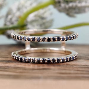 Könnte beinhalten: Ein goldfarbener Doppelring mit einem Band aus kleinen schwarzen Steinen an jedem Ring. Die Ringe sind durch einen goldfarbenen Steg verbunden. Der Ring liegt auf einer Holzoberfläche mit verschwommenem floralen Hintergrund.
