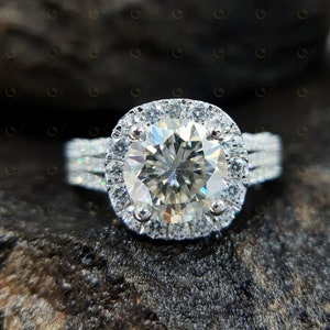 May include: A silver engagement ring featuring a large, round, clear diamond centre stone encircled by a halo of smaller diamonds. The band has three rows of smaller diamonds.