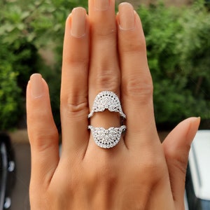 May include: A silver ring with a split design, featuring two half-circles of pave-set diamonds. The ring is worn on a person's finger.