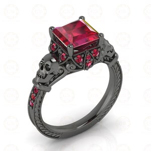 May include: A black metal ring featuring a large square-cut red gemstone at the center, surrounded by smaller red stones. The band is decorated with two sculpted skull motifs on the sides and intricate engraved patterns along the entire shank.
