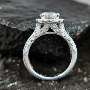 May include: A silver engagement ring featuring a large, round, clear centre stone surrounded by smaller stones. The band is also set with smaller stones and has a delicate, intricate design.