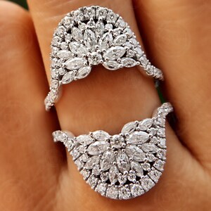 May include: Two silver rings with a design of small, clear diamonds set in a flower-like pattern. The rings are worn on a finger.