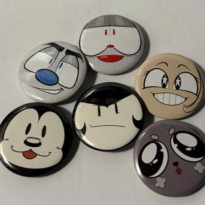 May include: A collection of six circular pin-back badges featuring various cartoon character faces. The designs include a white face with blue eyes, a grey face with red eyes, a tan face with star-shaped pupils, a black and white dog face, a pale face with dark hair, and a grey face with large eyes.