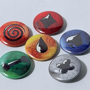 Puede incluir: Un conjunto de cinco chapas redondas con diseños gráficos distintos: una espiral roja, un diamante rojo, una gota de agua amarilla y naranja, una pica azul, un hueso verde y una forma de trébol de cuatro hojas gris, todos con acabado brillante.
